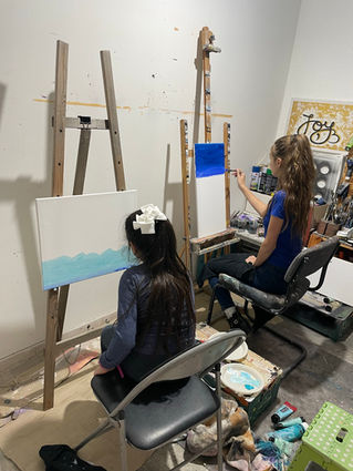 Two people painting on easels in an art studio, with one painting a blue sky and the other working on a blank canvas
