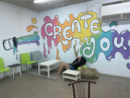 Artist lounging in front of a vibrant mural that says 'CREATE YOU' in colorful dripping paint