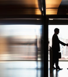 Two business professionals shake hands in silhouette at the entrance of a modern office building, with natural light streaming through the glass doors behind them. One person is holding a briefcase. The scene conveys a formal, professional interaction such as a meeting, partnership, or agreement.