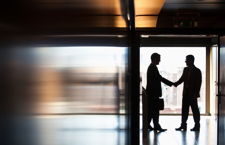 Two business professionals shake hands in silhouette at the entrance of a modern office building, with natural light streaming through the glass doors behind them. One person is holding a briefcase. The scene conveys a formal, professional interaction such as a meeting, partnership, or agreement.
