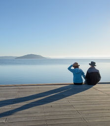 Two people sit on a wooden deck, gazing at a calm blue lake and distant island. They wear hats, casting long shadows in the serene setting.