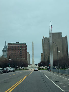 Street view of McKinley Monument in Buffalo, NY with a residential building, a high-rise building, and a church in the background.