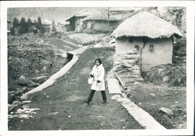 Woman walking on a path in front of traditional houses, Soon-Young Yoon.