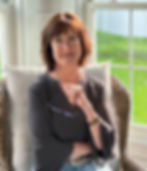 Older woman with brown hair smiling, holding a pair of glasses, exuding kindness, youth, and compassion.