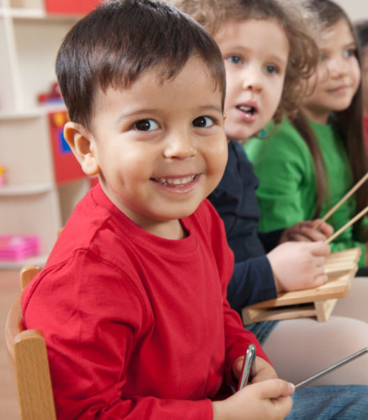 a child in a red shirt holding a triangle music instrument and smiling toward the camera with 2 children sitting to his side.