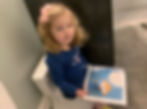 Young girl with a pink bow reads a colorful social story about potty training while sitting on a toilet in a bathroom, looking content.