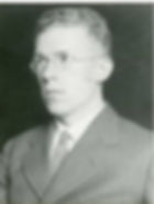 Black and white photo of a man in glasses and a suit with a striped tie, set against a dark background. He appears serious, looking to the side.