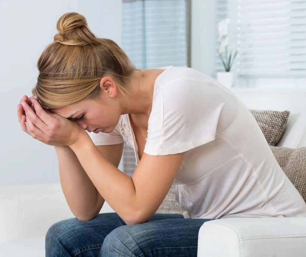 A woman in a white shirt and jeans sits on a couch, head in hands, appearing upset. Background features soft colors and indoor decor.