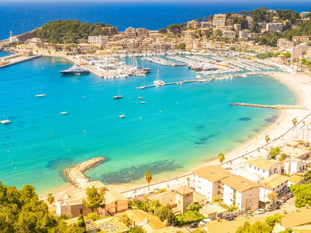 Vista panorámica del puerto de Sóller, con la bahía de aguas turquesas, embarcaciones fondeadas, la playa curvada y las casas junto al paseo marítimo bajo un día soleado.