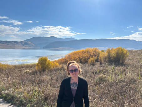 Julia, a red haired women with a pony tail and sunglasses is in front of a small lake and mountains in the sun