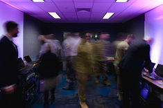 Long exposure photo at a defence event showing multiple people walking across a room, with coding visuals in the background, conveying a busy, dynamic atmosphere
