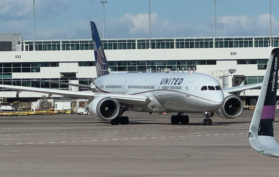 United jet taxiing on airport tarmac in front of terminal building, with another aircraft tail at right and cloudy sky overhead