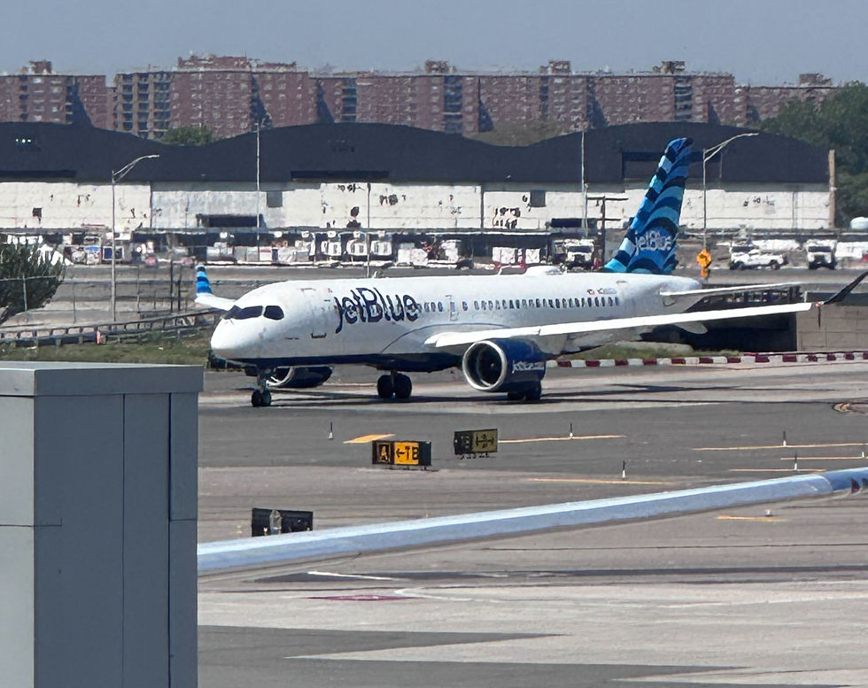 JetBlue airplane taxiing on airport runway, blue tail and engines, with terminal buildings and city apartments behind.