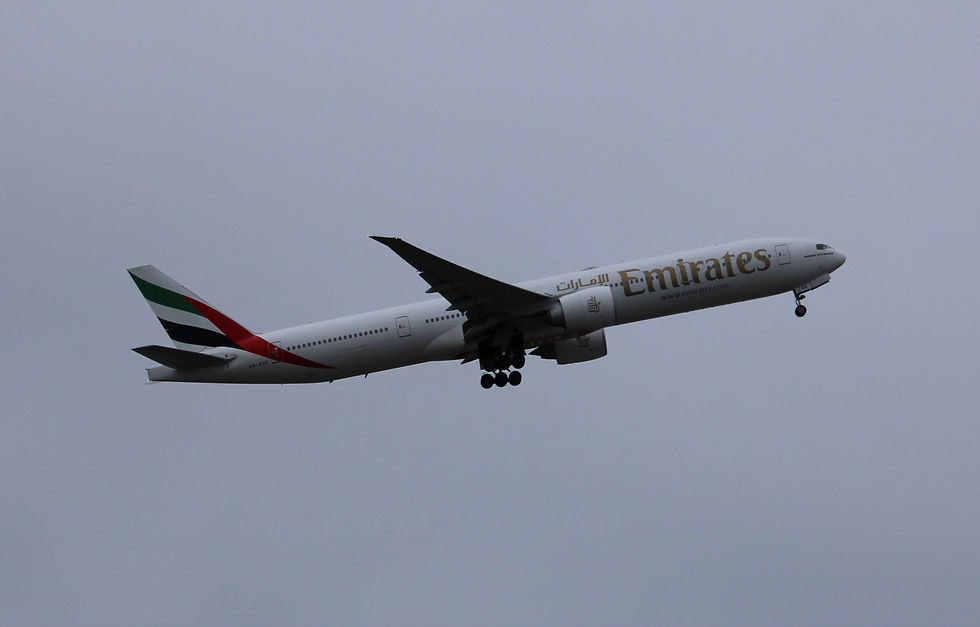 Emirates jet with landing gear down climbing against a gray cloudy sky.