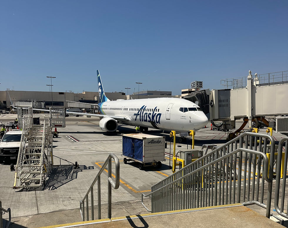 Airplane parked at a gate marked 63B in an airport. "Alaska" visible on the plane, with ground crew and luggage carts nearby on a sunny day.
