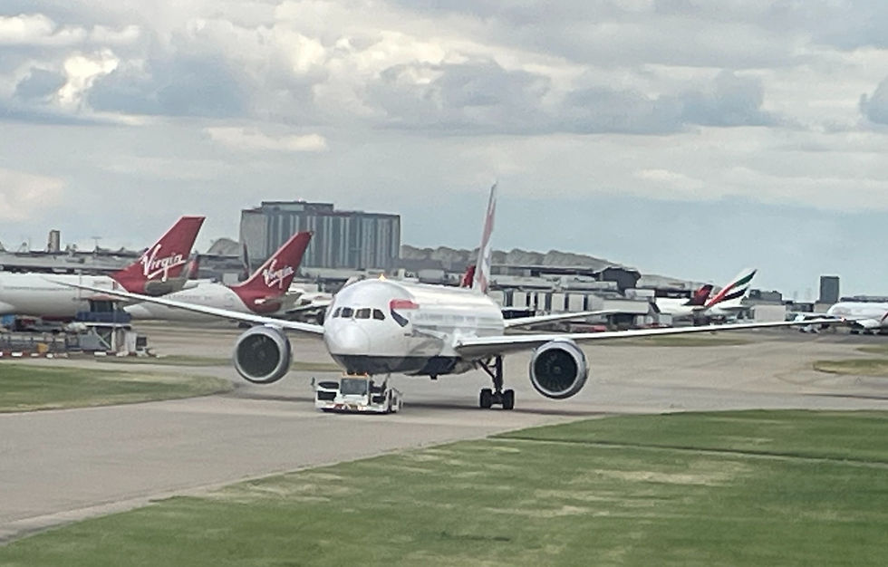 Emirates jet being towed on an airport taxiway, with Virgin planes in the background under cloudy skies.
