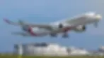 Emirates plane taking off against a clear sky, with airport buildings in the background. The aircraft is white with red and green accents.