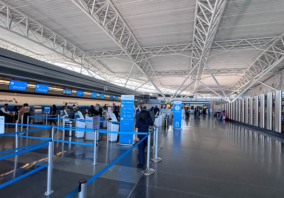 Travelers line up at American Airlines self-service bag drop in a bright airport terminal with blue signs and a high white truss ceiling.