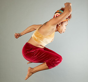 Orisha Re' executing a dance tuck jump with her knees drawn tight to her chest. Her right arm is pumping high and her left arm is low.