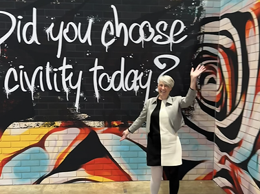 Melissa smiling and waving next to a mural that says "Did you choose civility today?"