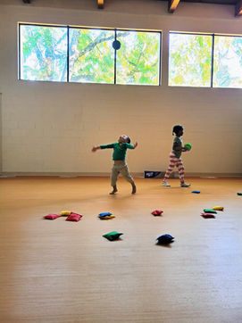 Watoto wakicheza na mifuko ya maharagwe ya rangi katika chumba cha mazoezi angavu, pana kilichojaa mwanga wa asili, wakitabasamu na kushiriki kikamilifu katika harakati na kucheza.