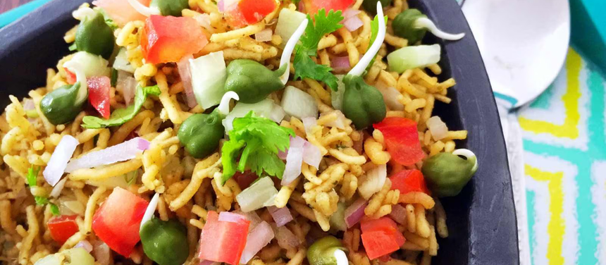 A bowl of colorful Bikaneri Bhujia chaat garnished with diced tomatoes, onions, green peas, sprouts, and fresh coriander served on a tray with a spoon.