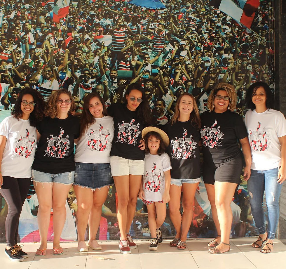 Mulheres e criança usando a Camisa Serpente do Movimento Coralinas em frente a mural de torcida na arquibancada.