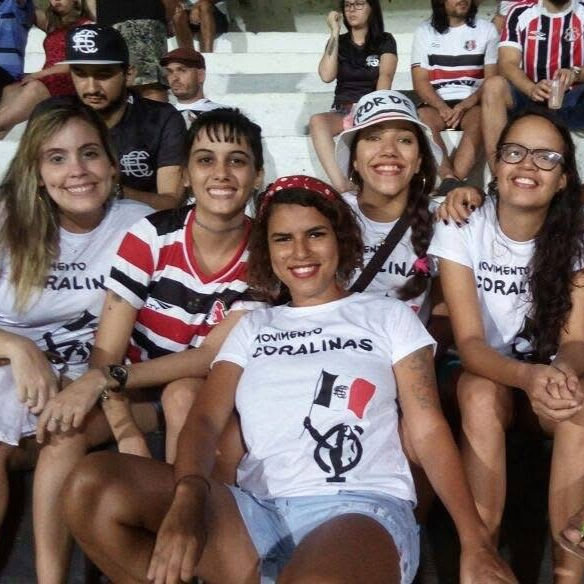 Mulheres usando a Camisa Bandeira do Movimento Coralinas em arquibancadas, com estampa de bandeira feminista.