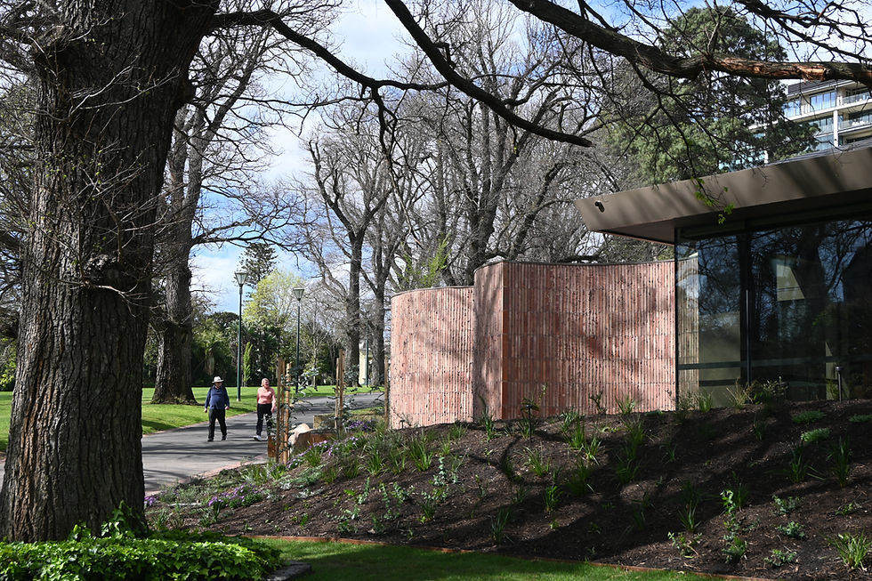 Two people stroll on a park path lined with bare trees. Brick building and greenery in the foreground. Sunny day with a clear sky.