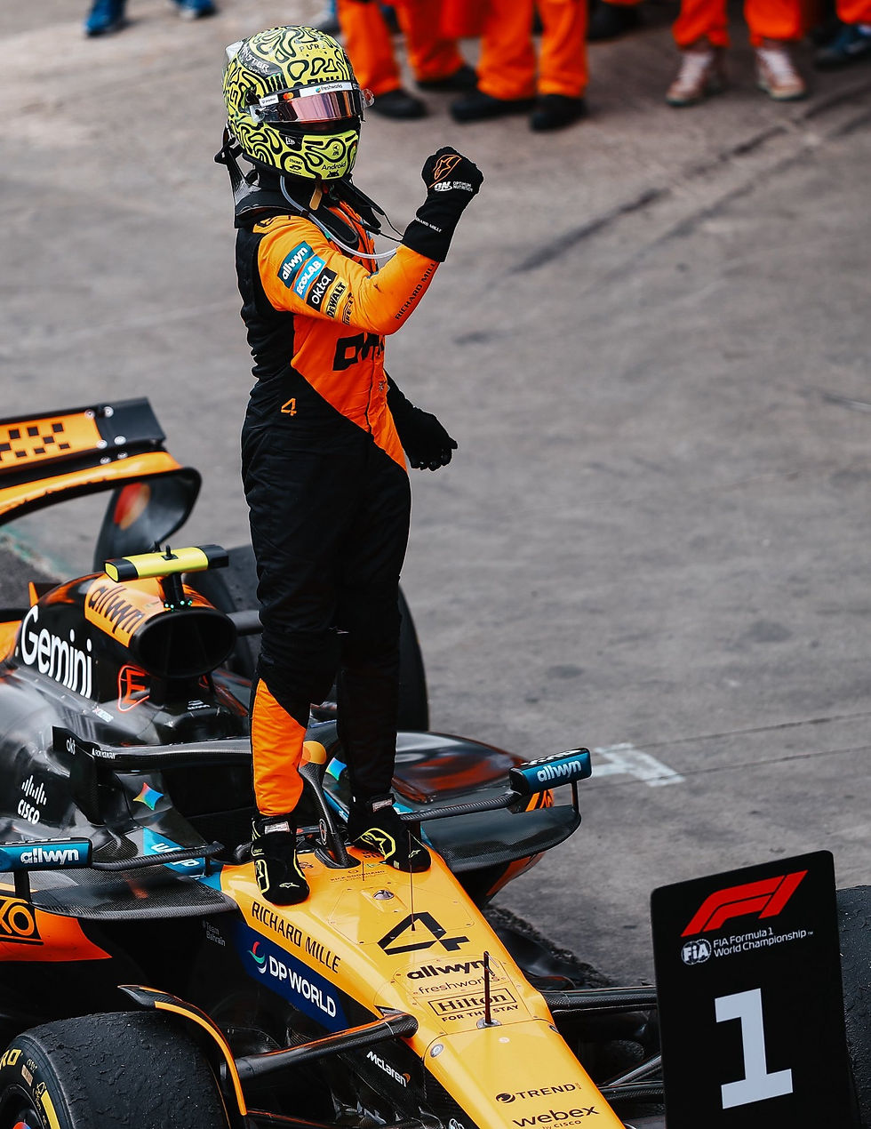 Lando Norris stands on his McLaren, clenching a fist in victory. A "1" sign is visible. Pit crew in background.
