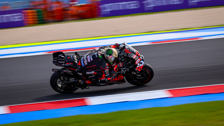 Motorcyclist in black and red gear speeds on a track, blurred background. "Aprilia" visible on bike, evoking a sense of speed and focus.