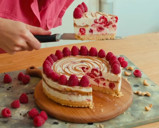 Hausgemachter Cheesecake mit frischen Himbeeren, servierfertig auf einem Holzbrett.