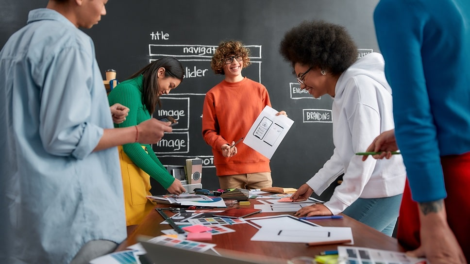 Diverse team brainstorms over sketches and color swatches in a studio, with a chalkboard labeled title, navigation, image, branding.