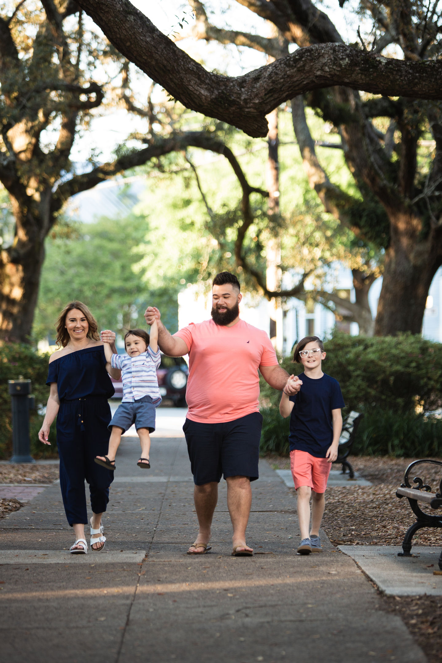 Family photography showing parents swinging toddler son by arms while walking in Seville Square with oak trees in background