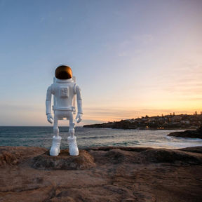 White and gold astronaut sculpture by New Zealand artist Milarky, from the NAUGHT series, standing on rocks at Sculpture by the Sea in Sydney overlooking the coast at sunset