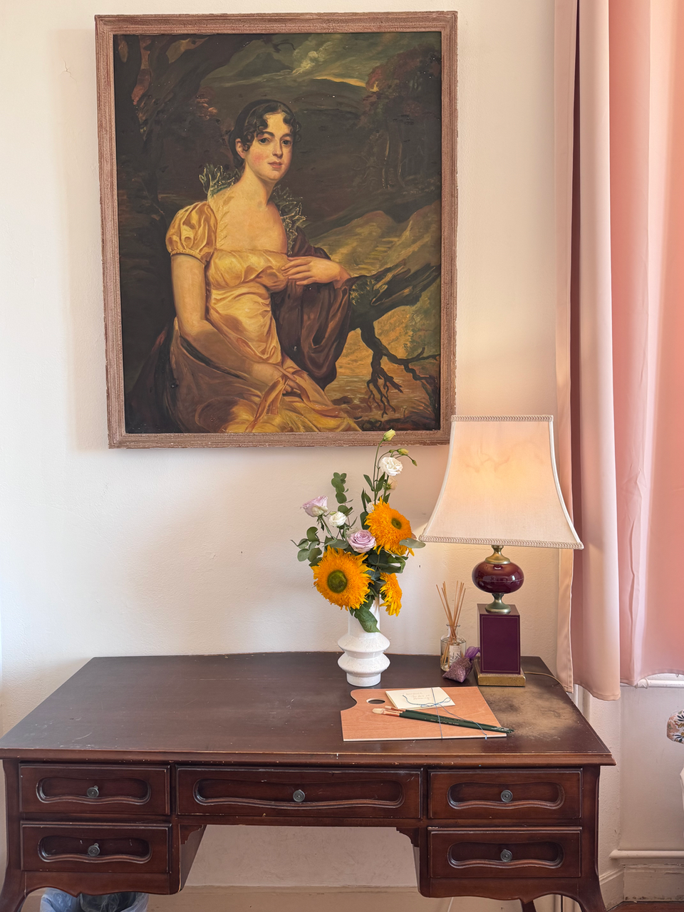 Elegant wooden desk with fresh flowers, lamp, and framed portrait painting.
