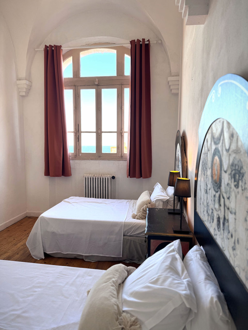 Charming bedroom with two beds, arched window, red curtains, wooden floor.