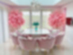 A dining room table dressed with pink scallop chairs and artificial pink plants in front of a large window dressed with Allusion blinds