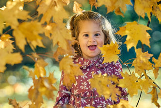 A creative, close-up portrait of a joyful young girl with her mouth open in excitement, framed through vibrant yellow maple leaves during an autumn family photo session in Scarsdale, NY 