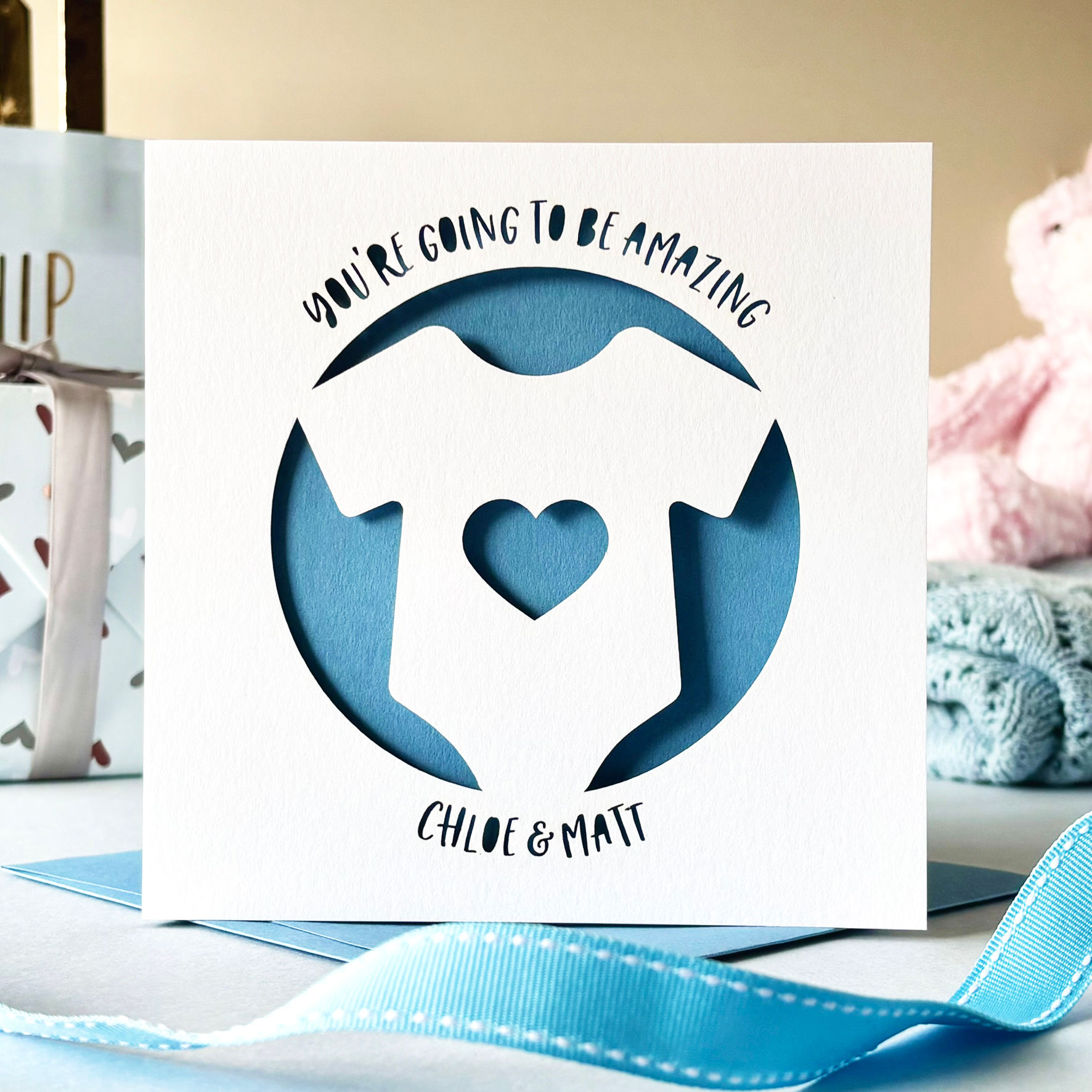 White laser cut personalised parents to be or baby shower card, featuring a babygrow with a heart, with a blue paper insert.