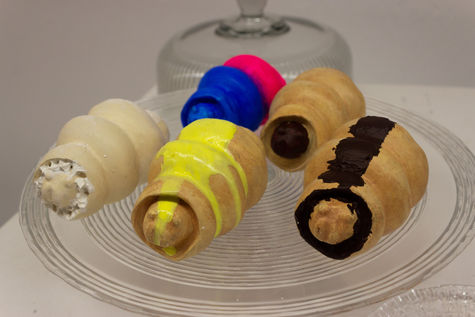 Image of five plaster casts of a maggot sculpture, each painted to look like a different food item. All five sculptures are pictured on a large glass cake tray  