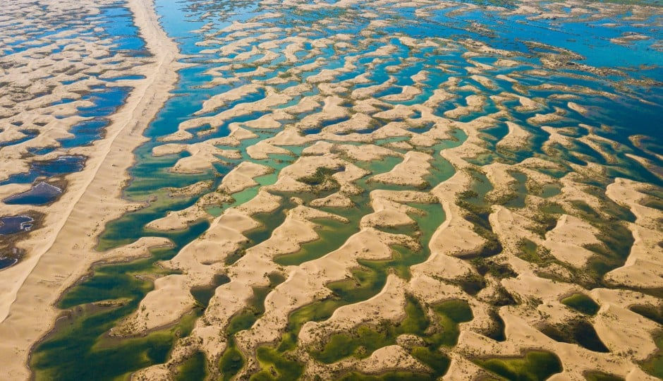Bu çarpıcı hava fotoğrafı, Kubuqi Çölü’nde oluşan su birikintileri ve kumul adacıklarının birbirine geçtiği yeni morfolojiyi sergileyerek restorasyon sonrası gelişen özgün hidro-ekolojik yapıyı gözler önüne sermektedir. Kum tepelerinin arasında tutulan yağış ve yüzey suları, zamanla küçük lagünler ve mikro sulak alanlar oluşturarak bölgenin su döngüsünü belirgin biçimde iyileştirmiştir. Bu karmaşık mozaik yapı, çöl ekosisteminin yeniden canlanma kapasitesini artırırken bitki, kuş ve diğer canlı türleri için kritik habitatlar meydana getirir.