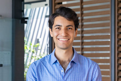 Hombre con camisa azul y pelo corto negro, posando de frente en una oficina moderna con vidrio y madera de fondo