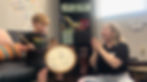 Alt Text: Photo of Isy, seated on the right of the frame, smiling and clapping to the beat. Isy is wearing a black t shirt, floral jeans, and a brown headband. A young music therapy
participant is seated beside her on the left of the frame. A strawberry blonde boy wearing a nirvana tshirt holds a frame drum in one hand and a drumstick in the other.
***Photo used with permission