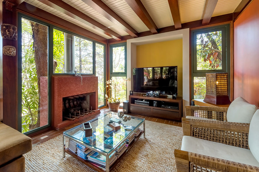 Sala de estar aconchegante com lareira avermelhada, teto com vigas de madeira e amplas janelas com vista para a natureza. Ambiente interno acolhedor desenvolvido por um Arquiteto em Atibaia, unindo rusticidade e sofisticação.