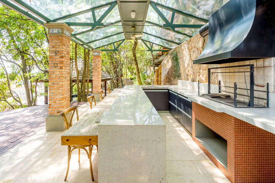 Área gourmet rústica-chic integrada à natureza com churrasqueira, balcão de granito, pilares de tijolos e cobertura de vidro. Exemplo de Projeto em Atibaia unindo conforto, funcionalidade e design orgânico ao ar livre