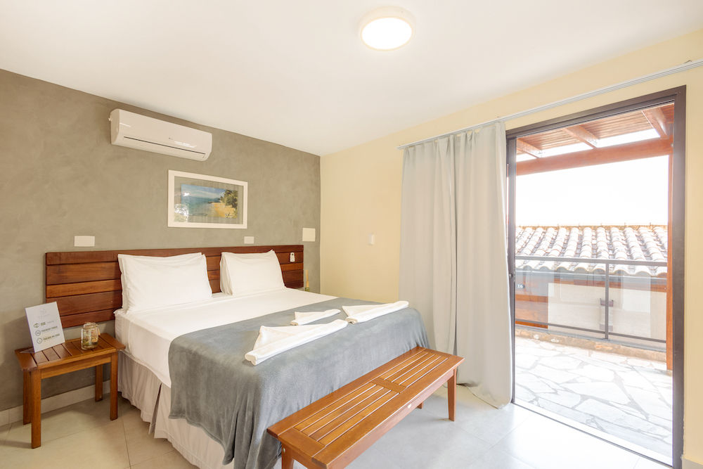 Suíte do Eco Baepi Residence com uma cama de casal, na parede ar condicionado, quadro com a paisagem da praia e varanda com uma vista para o mar e a serra de Ilhabela