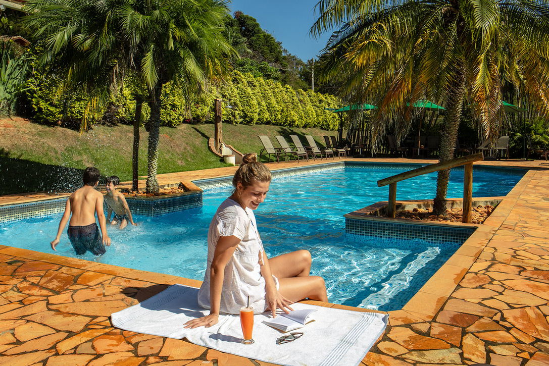 Mulher feliz tomando suco e lendo um livro enquanto duas crianças se divertem na piscina do Ibiti Hotel Rural