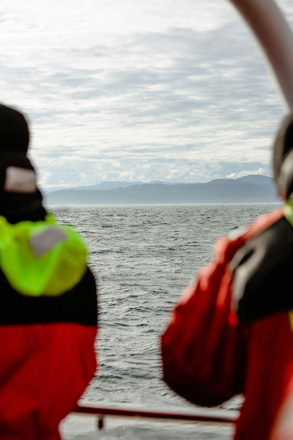 Two crew members in high-visibility gear looking out over the open ocean toward distant Alaskan mountains under a cloudy sky.