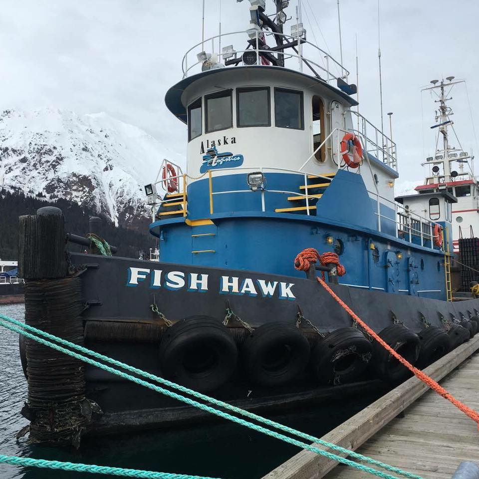 Alaska Logistics’ Fish Hawk tugboat docked in a harbor with snow-covered mountains in the background.